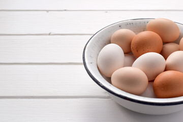 Assorted chicken eggs in different colors placed inside a vintage enamel bowl on a white wooden surface with copy space. Farm fresh, rustic and organic food concept