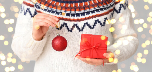 Woman in stylish warm sweater holding Christmas gift and ball on grey background, closeup