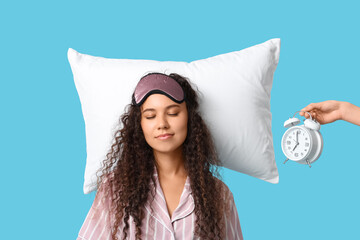 Beautiful young African-American woman with pillow and hand holding alarm clock on blue background