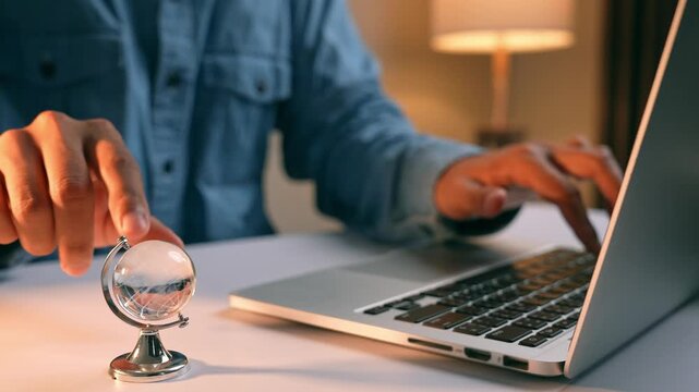 A hand rotates a desktop globe as the other operates a laptop, illustrating worldwide reach, international business, digital transformation, learning without borders, and curiosity.
