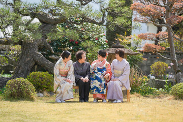 Four women in traditional Japanese clothing are sitting on a bench in a park. Scene is peaceful and serene, as the women are enjoying each other's company in a beautiful outdoor setting