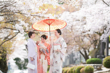 A group of women in traditional Japanese clothing are standing under an umbrella, with one of them holding a baby. The scene has a peaceful and serene mood