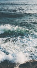 Fototapeta premium Seascape view features frothy waves crashing onto a dark shoreline. White foam contrasts the deep blue and turquoise of the ocean water under soft daylight