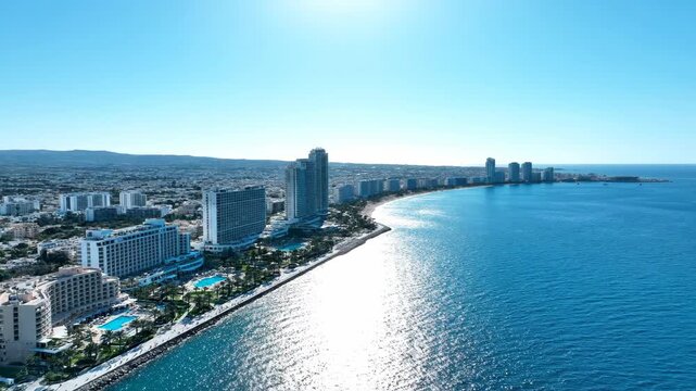 Aerial view of a modern coastal city with high-rise buildings lining a long beach and the clear blue Mediterranean Sea under a bright sky.
