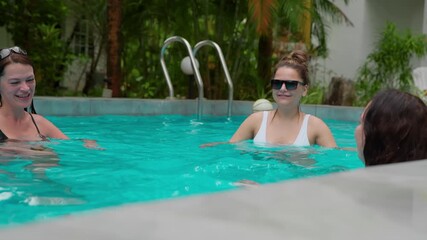 Cheerful women enjoying summer vacation, playing with inflatable ball and sharing joyful moments in sparkling swimming pool during leisure time at tropical resort