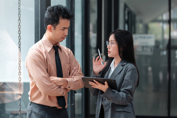 Businesswoman holding a tablet, discussing project details with a businessman, arms crossed, in a modern office setting