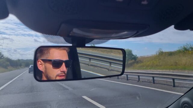 Rearview mirror view of a stylish man in sunglasses, concentrating as he drives down a sunlit highway on a bright summer day in the United Kingdom