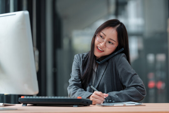 Asian businesswoman talking on phone and taking notes while working on computer in modern office - Powered by Adobe