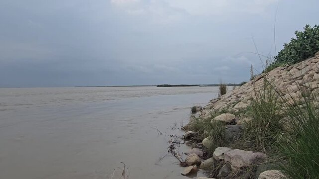 Wide muddy river landscape under overcast sky, rocky riverbank with green grass, calm brown water flowing through rural natural environment