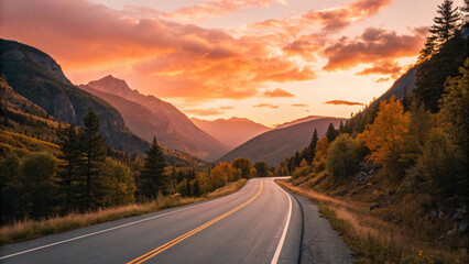 Naklejka premium Winding asphalt road through autumn valley with golden trees leading toward layered mountain ranges at sunset.