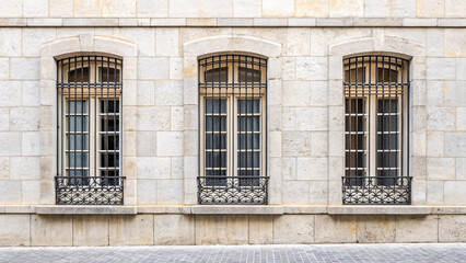 Fototapeta premium Three tall arched windows with decorative iron balconies on cream stone building facade, classical European architecture with grid pattern glass.
