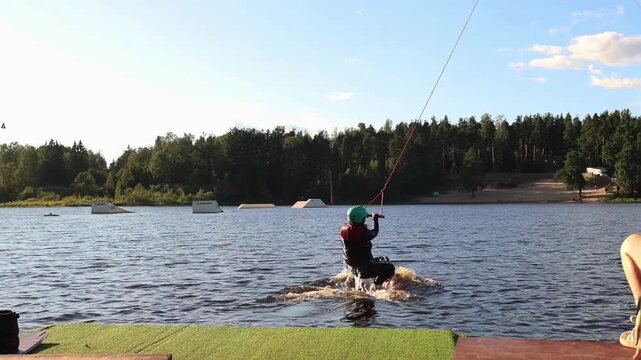 Wakeboarder starts ride on lake with cable system, splashing water under bright summer sky. Extreme water sport activity surrounded by forest and nature. Active woman on vacation, extrim hobby.