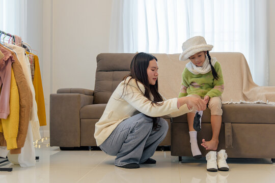 On a bright winter day a woman helps a girl with winter clothes, preparing her with a hat, scarf and winter boots, for a walk outside in the cold weather