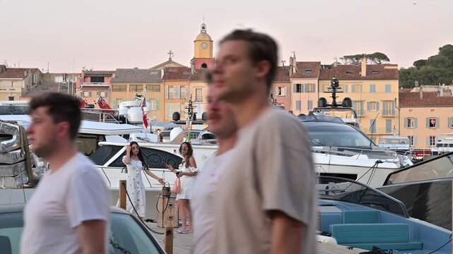 Saint Tropez harbor with luxury yachts and the bell tower of Notre-Dame-de-l&rsquo;Assomption, lively waterfront with unrecognizable passersby.