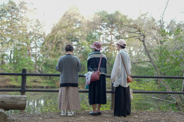 Three women are standing on a wooden bridge overlooking a pond. They are wearing traditional clothing and are looking at something in the water