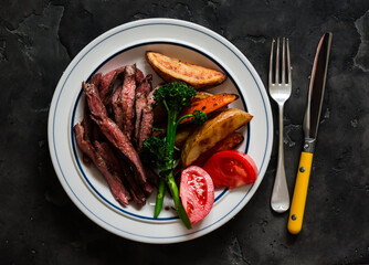 Delicious lunch, dinner - beef steak, baked potatoes, broccoli on a plate on a dark background, top view