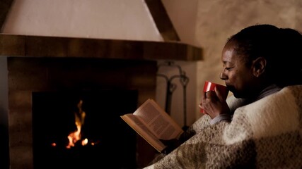 Afro latina woman reading book and drinking hot tea by fireplace - Winter christmas holiday home relaxation - Powered by Adobe