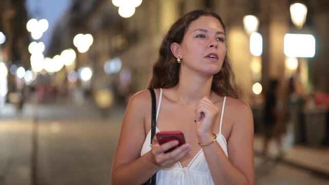Young woman with a mobile phone in hands walks down an evening street in Barcelona, typing messages and looking at restaurant addresses in the app. Nightlife and parties in summer. High quality 4k