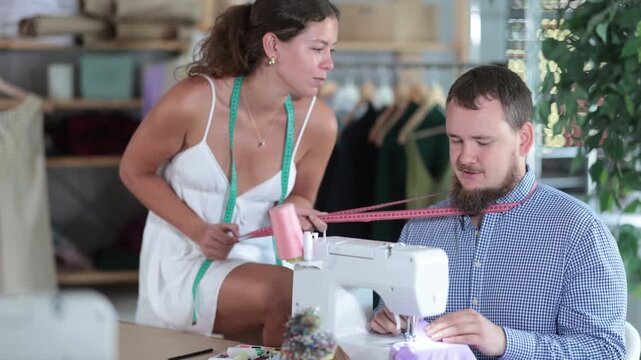 Playful young woman harasses male tailor working at a sewing machine in fashion studio. High quality 4k footage