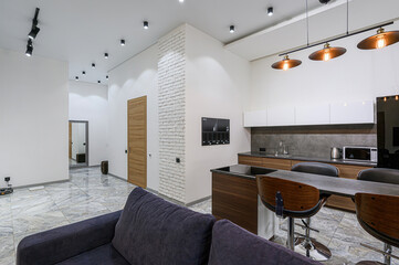 Modern interior with white walls, a kitchen featuring pendant lights, a wooden door, and marble floors.  A blue sofa is in the foreground