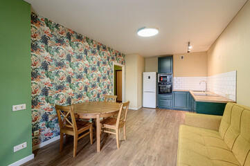 cozy apartment interior features a round wooden table with chairs, a green wall, a wallpapered wall, a yellow couch, and a kitchen area with green cabinets