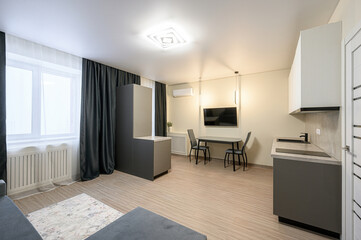 modern, well-lit apartment with neutral tones. Features include a dining table, TV, kitchen area, and ample natural light through a large window