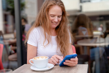 Woman with long hair smiles while using her smartphone at a cafe during a sunny afternoon, enjoying a coffee in a lively outdoor setting