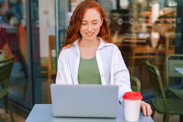 Young woman working on laptop at outdoor cafe enjoying coffee while smiling in bright daylight in a lively atmosphere