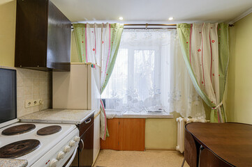 cozy kitchen features a white stove, dark brown cabinets, a refrigerator, and a window with light, floral curtains. A wooden table sits in the foreground