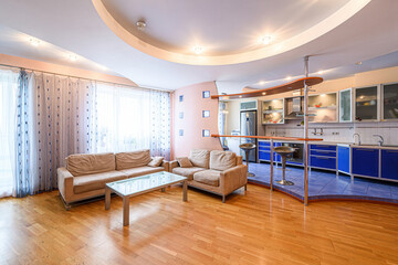 bright living room and kitchen area feature tan sofas, a glass table, hardwood floors, blue kitchen cabinets, and a unique ceiling design with recessed lighting