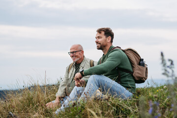 Adult son and older father hiking togeter in nature, resting on meadow.