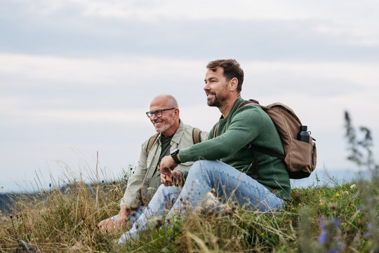 Adult son and older father hiking togeter in nature, resting on meadow.