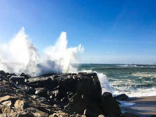 Powerful scene of Atlantic Ocean waves crashing against rocky shore in Porto, Portugal. The spray of water rises high into the blue sky, capturing the raw power of nature on the Portuguese coast.