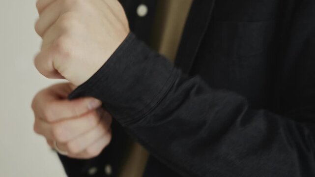 Close-up shot of male hands fastening the cuff of a black shirt in a bright room. Frame emphasizes daily self-care, morning preparation, and personal dressing rituals. High quality 4k footage