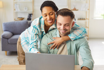 Young cheerful diverse couple sitting at desk in living room at home together watching video or using laptop for internet and social media. Happy man and woman resting enjoying weekend.