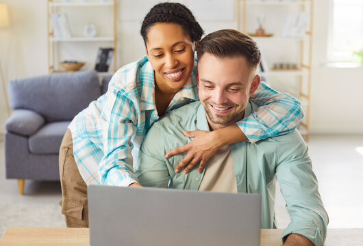 Young cheerful diverse couple sitting at desk in living room at home together watching video or using laptop for internet and social media. Happy man and woman resting enjoying weekend. - Powered by Adobe