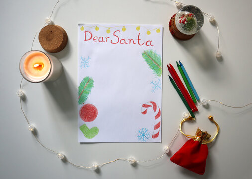 Children's drawing of Dear Santa letter with drawings of fir branches, Christmas ornaments on white surface. Pencils, candle, snow globe, gift bag, string lights are arranged nearby, cozy atmosphere.