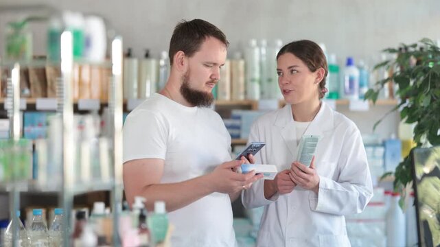  Young man buyer scanning qr code for box of ointment with young woman pharmacist in pharmacy