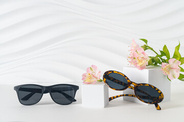Two pairs of Trendy sunglasses on podiums on white background with flower. New eyewear collection....