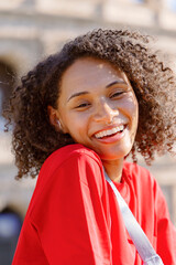 A lively young woman with curly hair radiates joy, smiling in a striking red shirt outdoors. The stunning historic architecture in the background enhances the beautiful scene perfectly