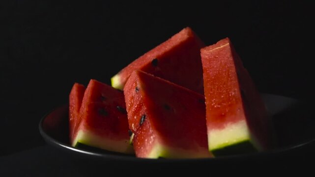 Close up zooming in video of gorgeous red colored and tasty delicious watermelon slices in triangle shaped with the green shell still on them while being put in front of a black studi background. Seed