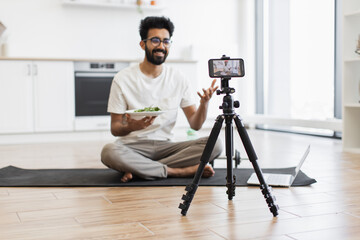 Young adult man sitting cross-legged on yoga mat in modern kitchen recording video blog with smartphone and tripod holding healthy salad plate. Scene suggests health, nutrition, video blogging.