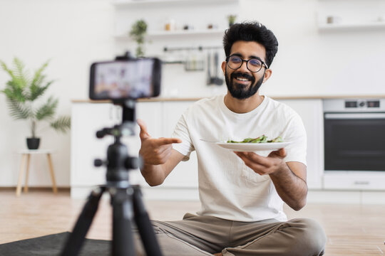 Man sitting cross-legged in kitchen filming a video about healthy eating for viewers. Includes indoor background, white shirt, home setting, casual atmosphere.