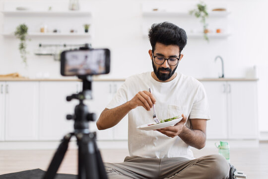 Middle age man sitting on yoga mat in bright kitchen recording healthy eating blog with smartphone focused on healthy lifestyle wellness capturing audience interest. Laptop food props enhance setup.