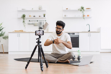 Caucasian adult man in casual outfit recording sports-related vlog discussing smart fitness devices. Room setting is a modern kitchen with exercise equipment visible.