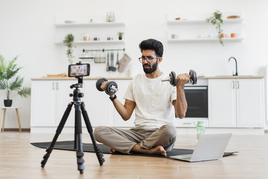 Man exercising with dumbbells during video recording in kitchen, showcasing fitness routine and health. Seated on mat near laptop and phone, promoting home fitness motivation.