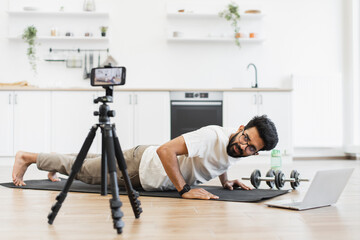 Young adult man wearing glasses and casual outfit does push-ups indoors filming video. Fitness enthusiast recording workout routine using smartphone and online support.