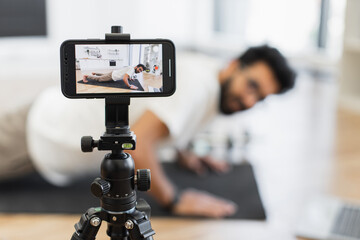 Young adult man doing push-ups on exercise mat, recording fitness video blog using smartphone in indoor setting. Promotes healthy lifestyle and home workouts. Emphasizes dedication to fitness, health