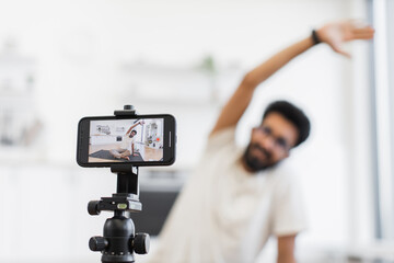 Young adult man records fitness tutorial at home using smartphone camera, focusing on arm stretches. Promotes health, fitness, and active lifestyle through shared video content on personal platforms