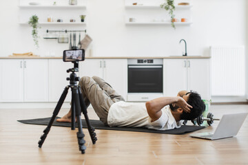 Man recording video blog on fitness and healthy living performing abs exercises, showcasing equipment, camera setup, modern kitchen environment for engaging content creation, features indoor workout